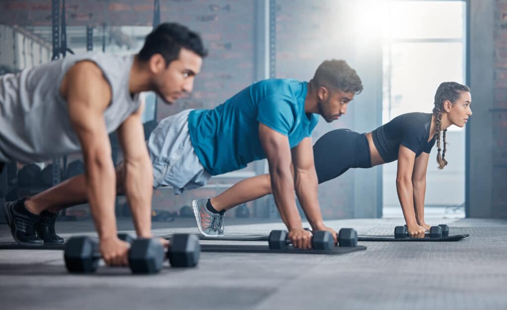 Trois personnes (deux hommes, une femme) font des planches avec haltères dans un gym moderne. Ils sont concentrés sur leur entraînement fitness.