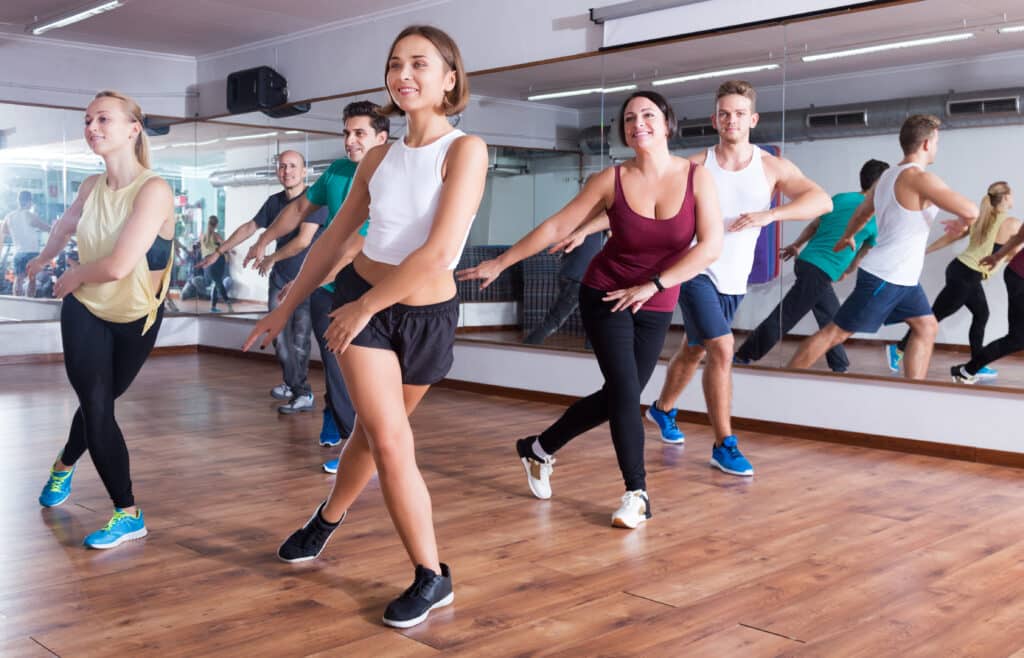 Groupe d'adultes souriants en plein cours de danse ou fitness dans un studio lumineux avec des miroirs, effectuant des mouvements.