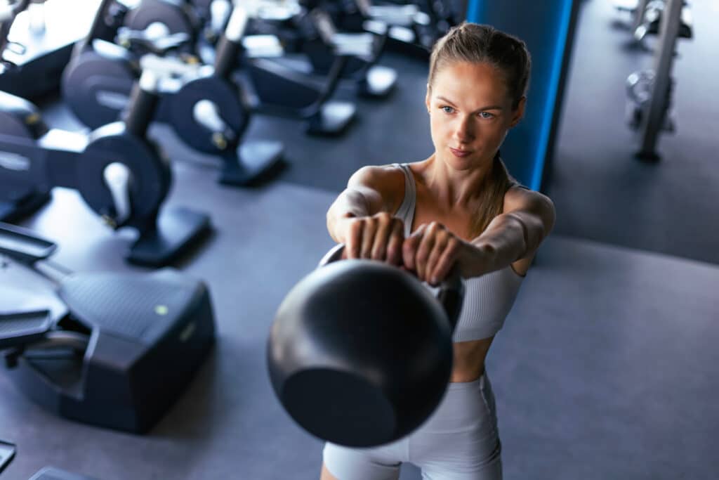 Femme en tenue de sport, concentrée, effectuant un entraînement au kettlebell noir dans un gym moderne et lumineux.
