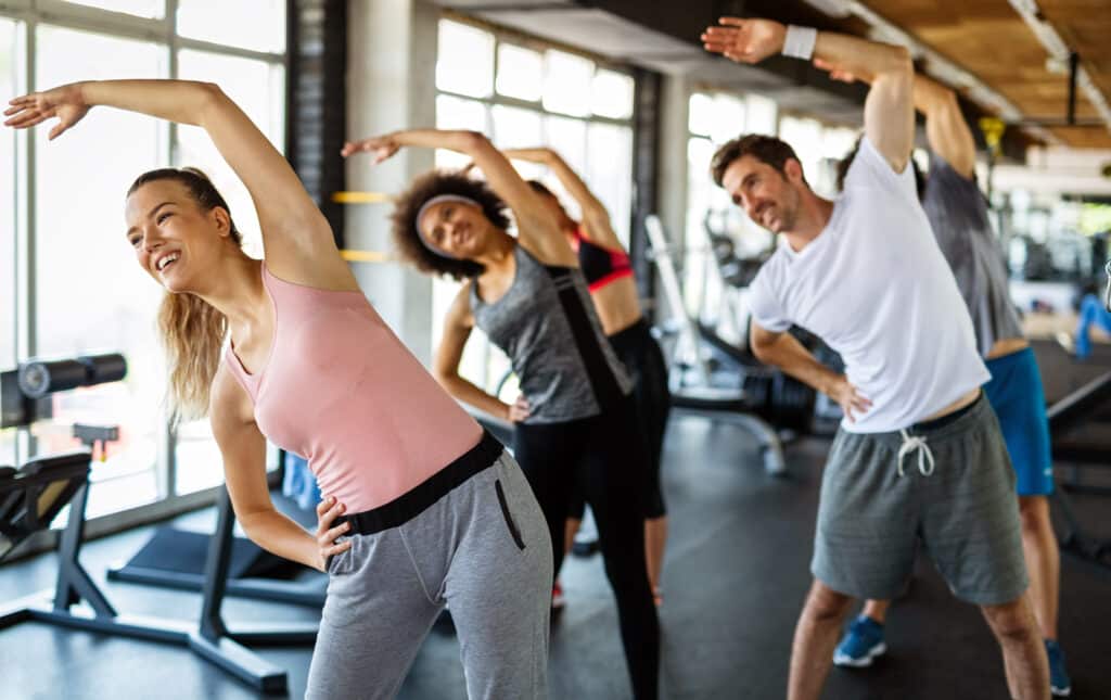 Un groupe de personnes souriantes étire son corps latéralement dans une salle de sport lumineuse, avec une femme au premier plan en haut rose.