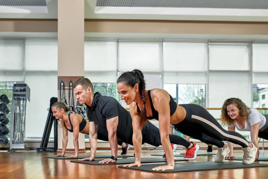 Quatre personnes en tenue de sport réalisant une planche sur des tapis de yoga dans une salle de sport, concentrées et souriantes.