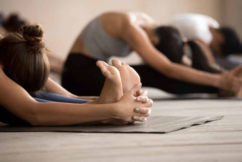 Femme étirée en posture de yoga Paschimottanasana, mains sur les pieds. Cours de yoga en groupe, ambiance sereine.